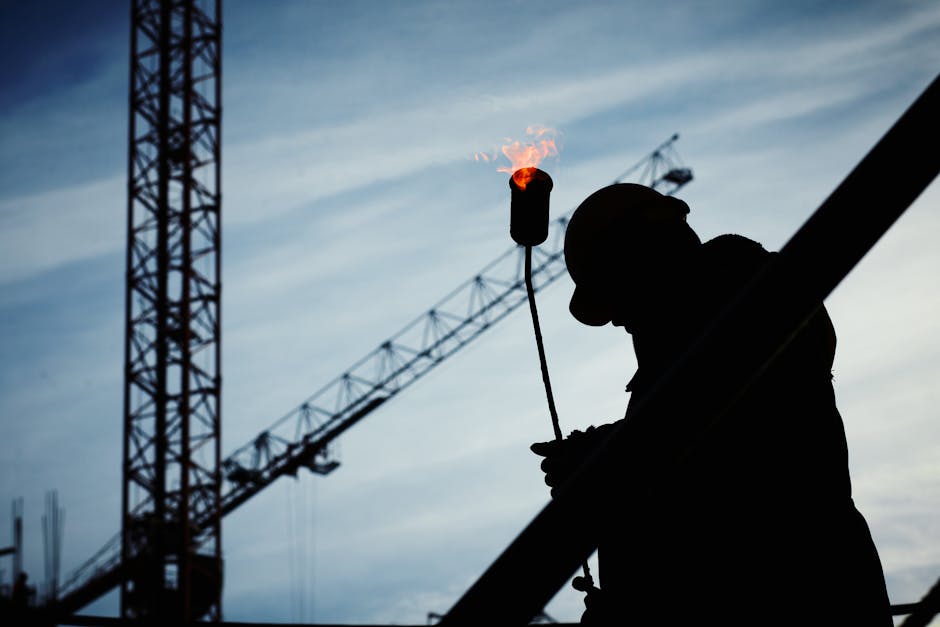 Silhouette of a construction worker using a blowtorch at a building site against a crane-filled skyline.